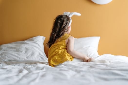 A young girl in bunny ears enjoys a cozy moment on a bed with a warm yellow background.