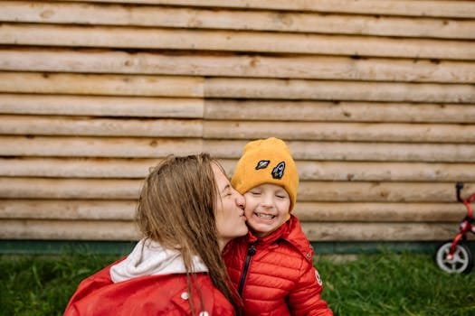 A mother lovingly kisses her smiling child in an outdoor setting, wearing red jackets.