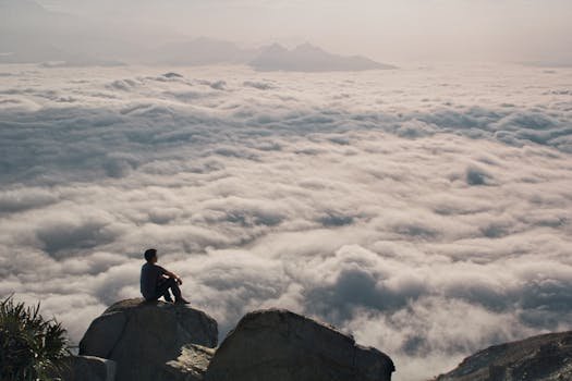 Person sitting on a mountain peak above clouds, capturing serenity and adventure.