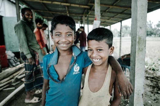 Two smiling children embraced under a rustic shelter in a rural outdoor setting, radiating joy and friendship.