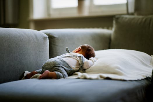 A cute baby in a gray outfit sleeps on a cozy sofa indoors, creating a calm and serene atmosphere.