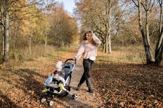 A mother walks with her child in a stroller along a leaf-covered path in a serene autumn park.