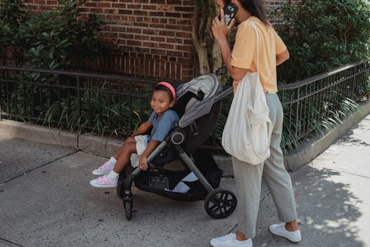 Crop Asian woman talking on smartphone while pushing baby stroller with little girl during walk on pavement in sunny city