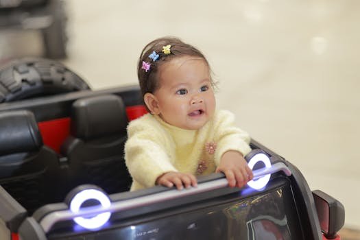 Cute baby in a toy car indoors, wearing colorful clips and a yellow sweater.