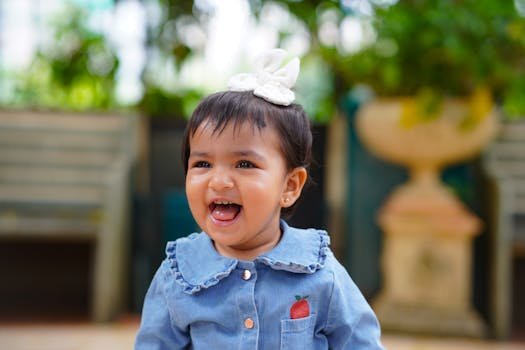 Cute baby girl smiling outdoors wearing a denim shirt with a bow in her hair.
