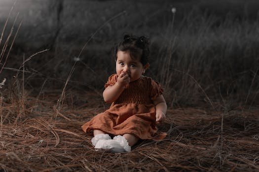 A cute baby sits on autumn leaves in a forest, wearing a brown dress, creating a serene portrait.