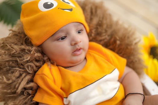 Charming baby wearing a bright orange outfit and hat, sitting on a furry cushion with a sunflower background.