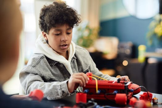 Concentrated ethnic kid with curly hair in warm jacket exploring new red device and sitting at table in light room