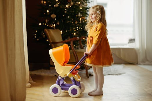 A young girl in an orange dress plays with a toy stroller by a Christmas tree indoors.