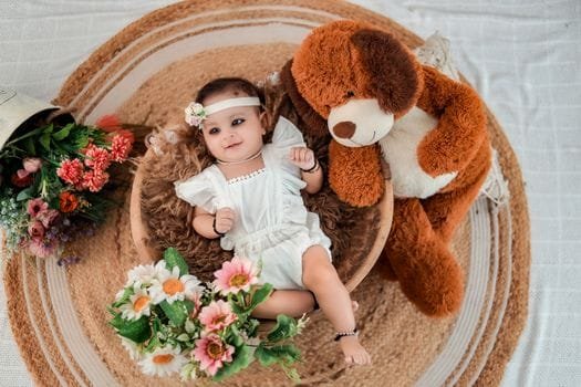 Cute baby laying by a teddy bear, surrounded by colorful flowers.