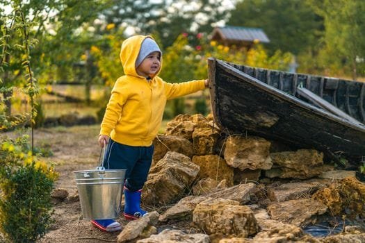 Young child in a yellow hoodie and beanie holds a metal bucket outdoors near an old boat and rocky terrain.
