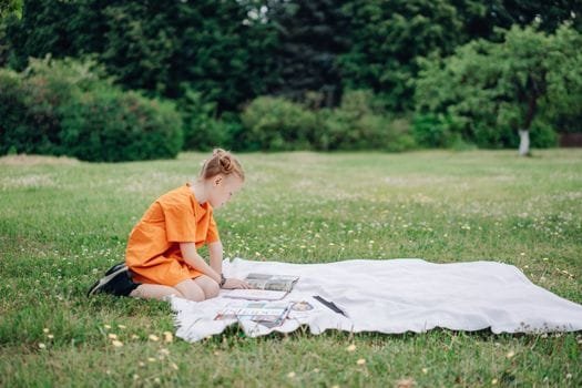 A young girl sits on a picnic blanket in a park, reading a book on a sunny day.