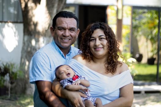 Smiling parents holding their baby in a sunlit outdoor setting.