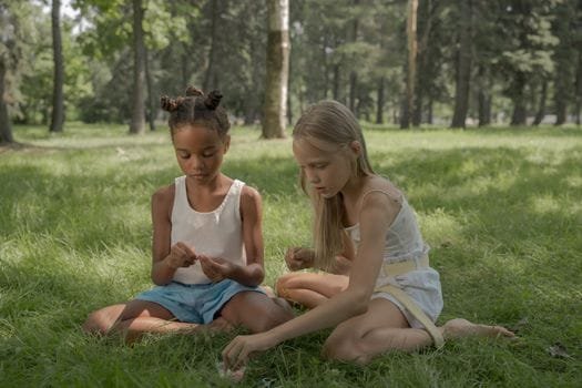 Two girls sitting and playing on grass in a sunny park.