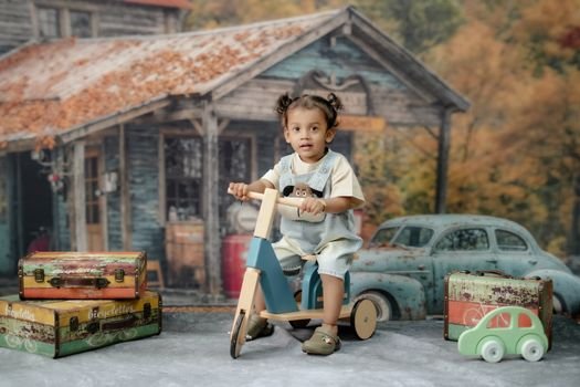 Adorable toddler rides a wooden bike in a charming vintage setting with an old car and suitcases.