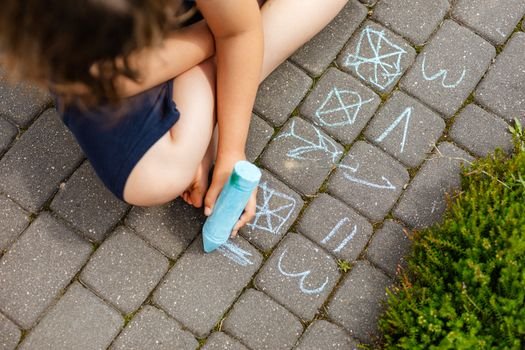 A child uses blue chalk to draw shapes and patterns on paved outdoor surface.