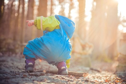 A young child exploring a sunlit forest in a vibrant blue and green raincoat.
