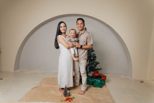 A smiling family poses with their baby and Christmas tree, surrounded by gifts indoors.
