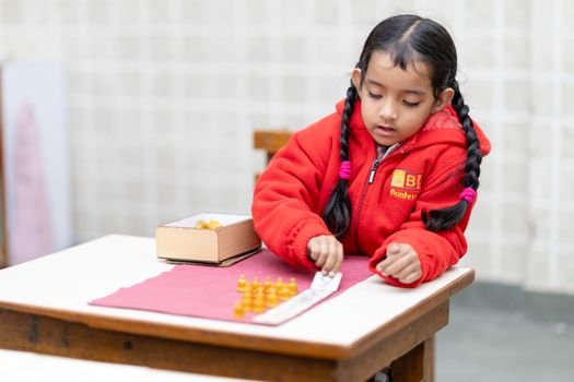 A young South Asian girl in a red jacket focuses on a learning task at school.