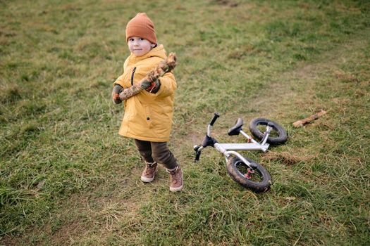 A child wearing a yellow jacket holding a stick in a grassy field near a toy bike.