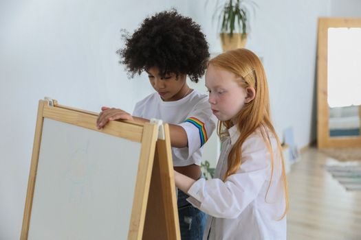 Side view of concentrated multiracial girls drawing on double sided easel chalkboard at home in daytime