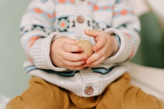 Close-up of a child's hands holding a wooden toy ball, wearing a cozy sweater indoors.
