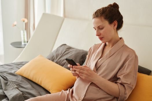 Pregnant woman wearing sleepwear, relaxing and using a smartphone in a cozy bedroom setting.