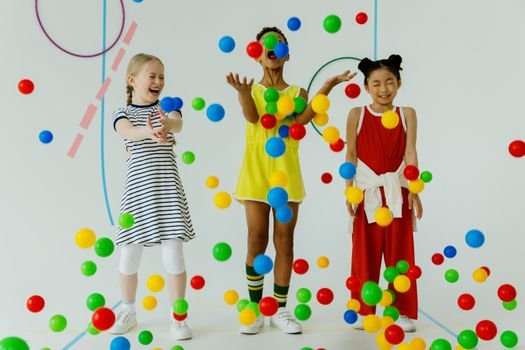 Three diverse girls laughing and playing with colorful balls indoors.