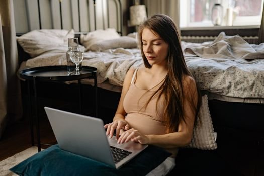 A pregnant woman sits on the floor using a laptop, symbolizing remote work and maternity.
