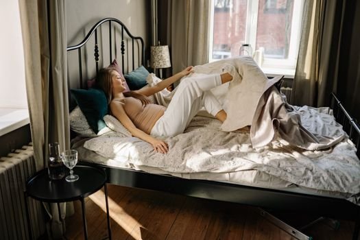 Pregnant woman comfortably lying in bed, enjoying morning sunlight in cozy bedroom setting.