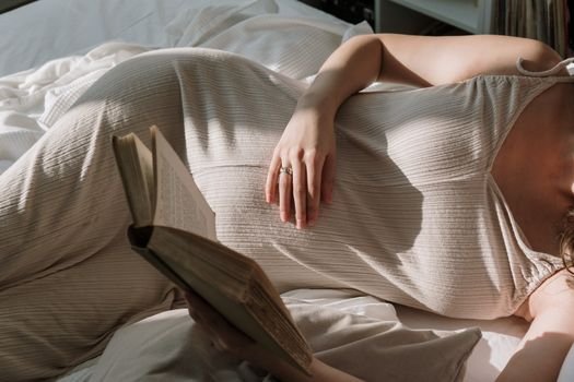 Pregnant woman with flowing dress reading a book in bed, enjoying peaceful leisure time.