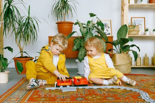 Cute siblings wearing yellow and white clothes sitting on colorful old fashioned carpet and having fun together while playing with plastic railway in living room