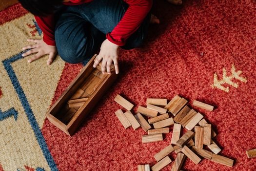 From above crop kid in casual clothes sitting on floor carpet while playing in jenga tower game with wooden blocks improving mental skills and accuracy