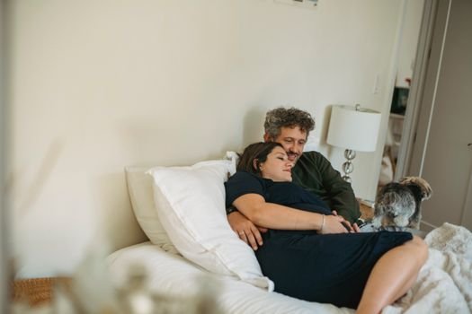 Smiling man cuddling gently expectant woman while lying on soft bed with cellphone near dog