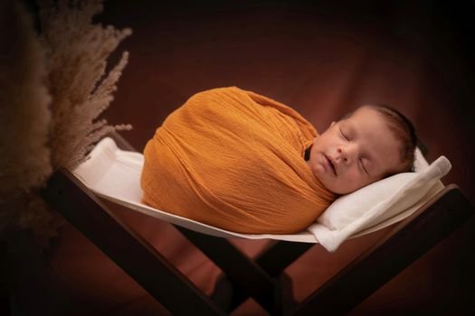 Adorable newborn baby peacefully sleeping in warm orange wrap on white cushioned basket.